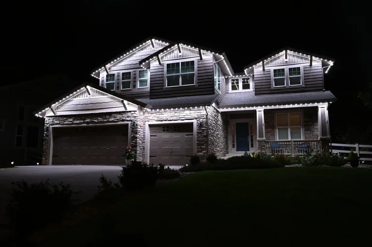 A modern two-story house decorated with white string lights outline on the roof and eaves, illuminated at night, with a front porch, stone facade, and two garage doors.