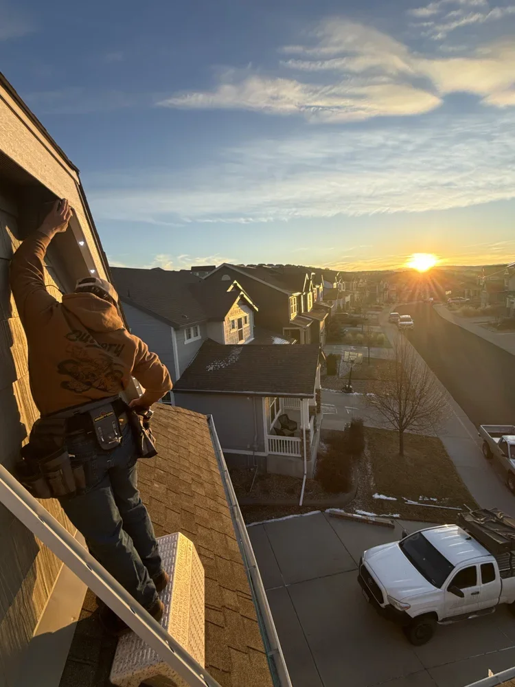 A worker in an orange hoodie with tools on his belt is standing on the roof of a house during sunset, with a neighborhood and a sunset in the background.