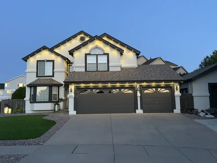 A two-story suburban house with gray garage doors, white exterior, and decorative string lights along the roofline, taken during twilight.
