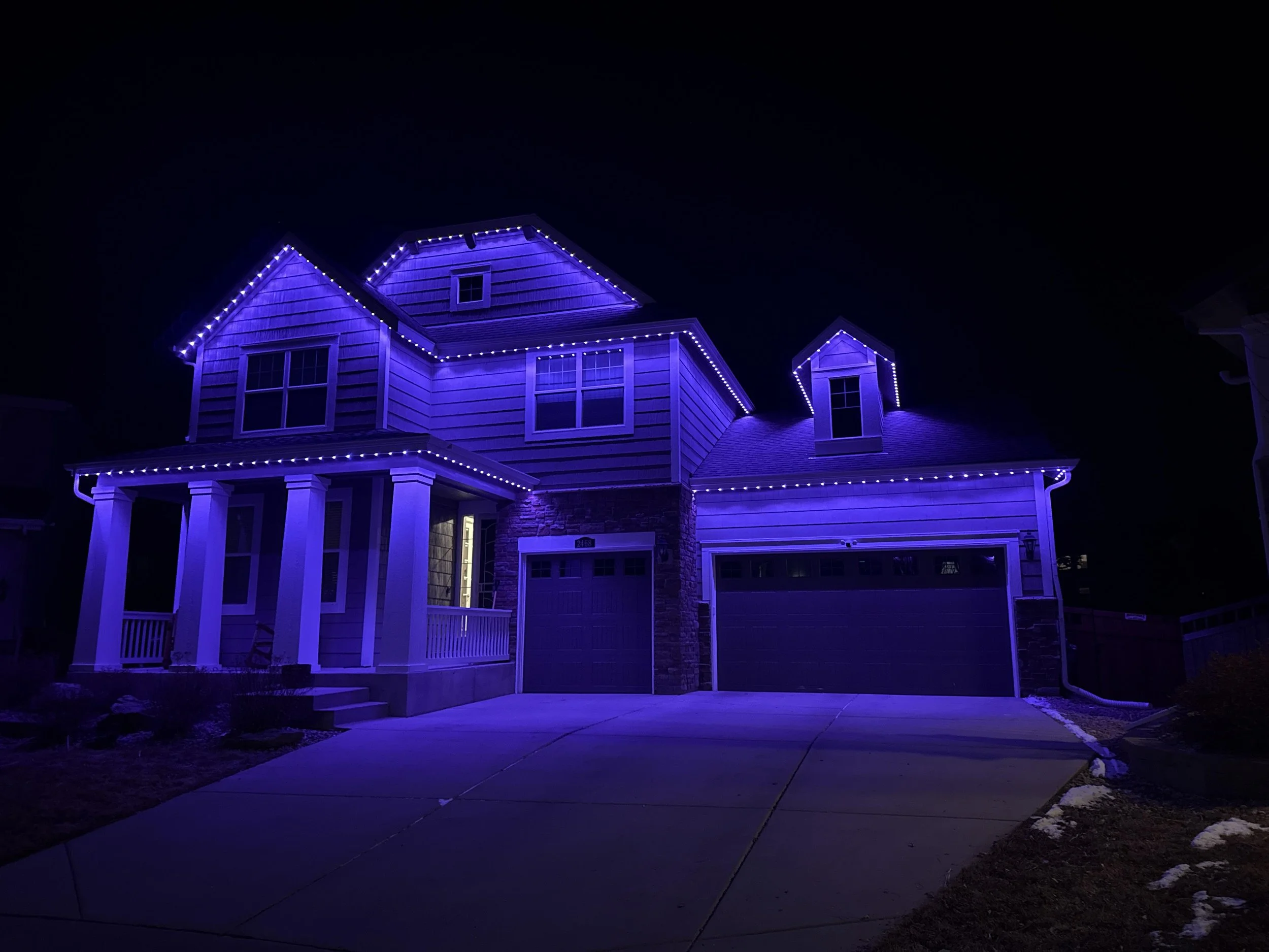 A two-story house illuminated with purple string lights outlining the roof, windows, and porch area at night. The house has white columns at the front and a garage to the right. There is a driveway in front of the house and some patches of snow on the grass.