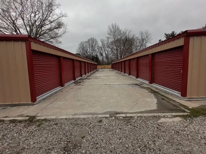 Row of storage units with closed red roll-up doors, concrete pavement, and leafless trees in the background under cloudy sky.