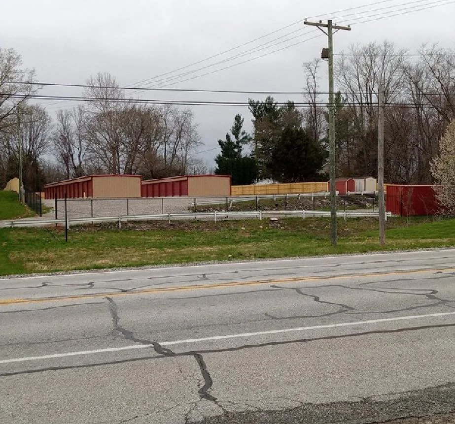 A street view showing a grassy area with a sidewalk, wooden utility poles, and a row of storage units or sheds in the background, with leafless trees and cloudy skies.