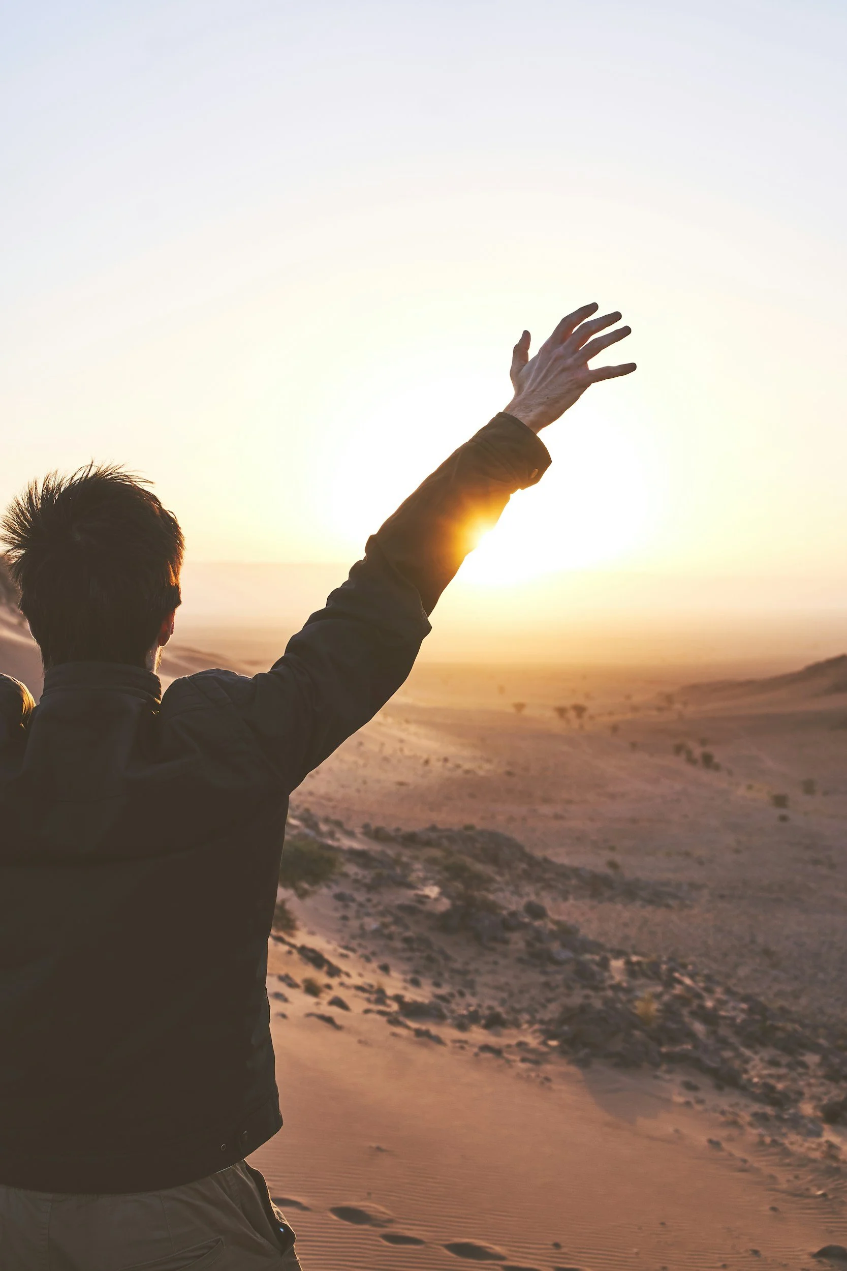 Person with dark hair and a brown jacket raising their hand in the air at sunset over a desert landscape.