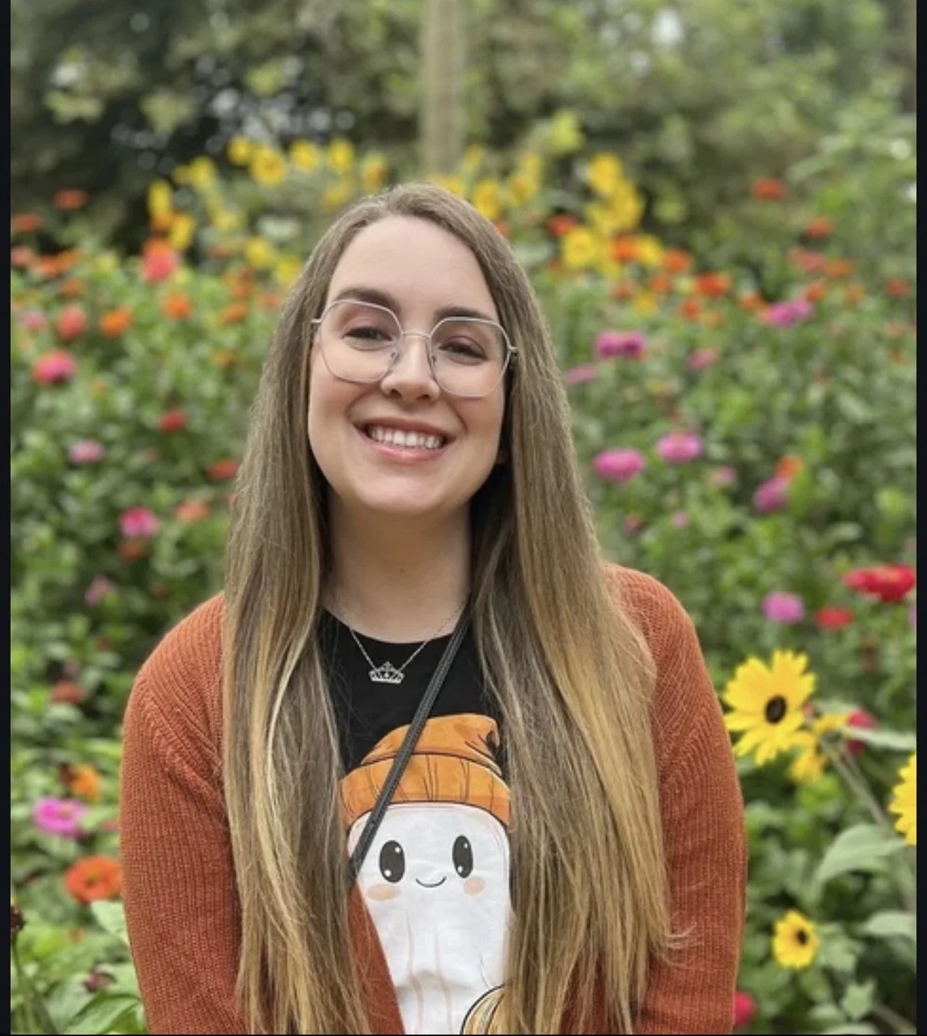 A young woman with long, light brown hair and glasses is smiling at the camera in a garden filled with colorful flowers, including sunflowers, pink, orange, and yellow blossoms. She is wearing a black t-shirt with a cartoon character and an orange cardigan, and a necklace with a crown pendant.