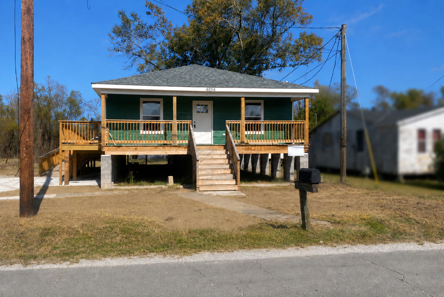 Front view of a house with a green exterior, elevated on stilts with a wooden staircase and balcony, in a suburban area with clear blue sky.