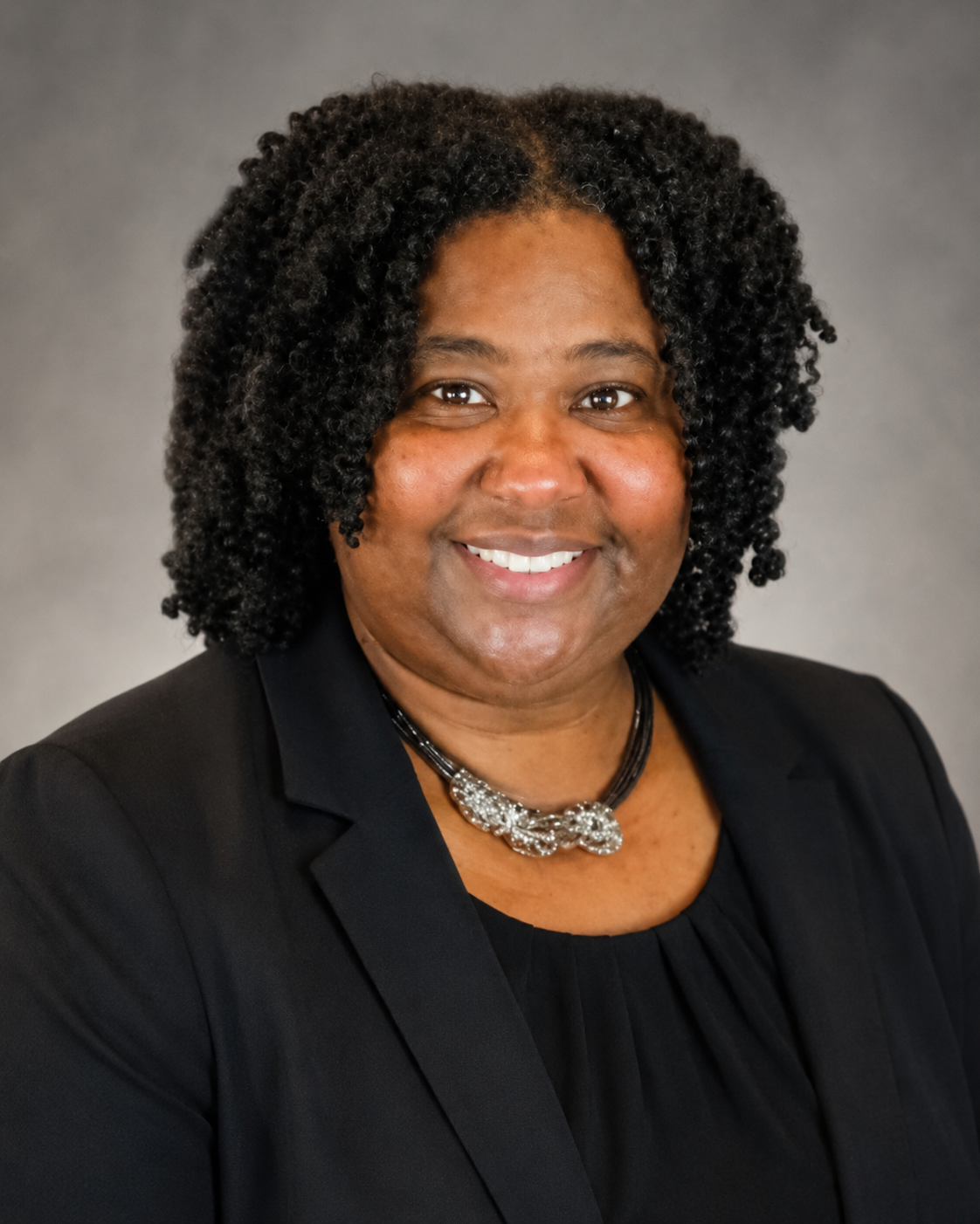 A professional woman with dark, curly hair wearing a black blazer and a decorative necklace, smiling at the camera against a gray background.