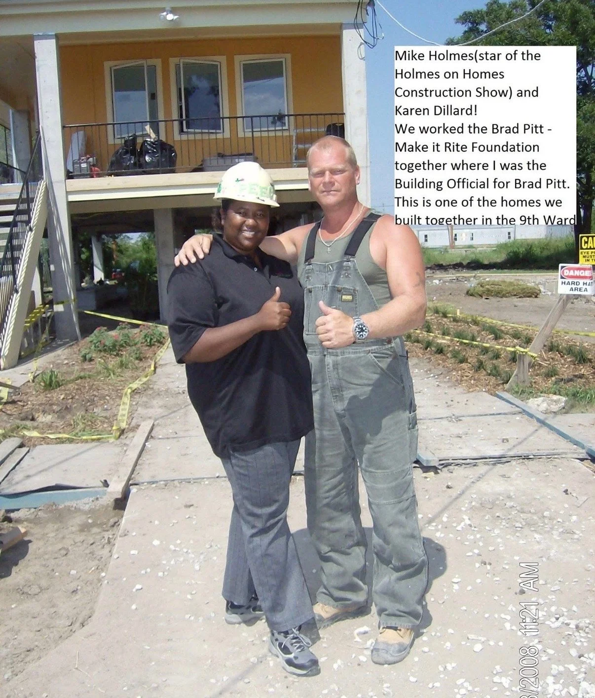 Two construction workers, a woman and a man, standing on a construction site in front of a partially built house. The woman is wearing a construction helmet and black shirt, and the man is in gray work overalls, both smiling and giving a thumbs-up.
