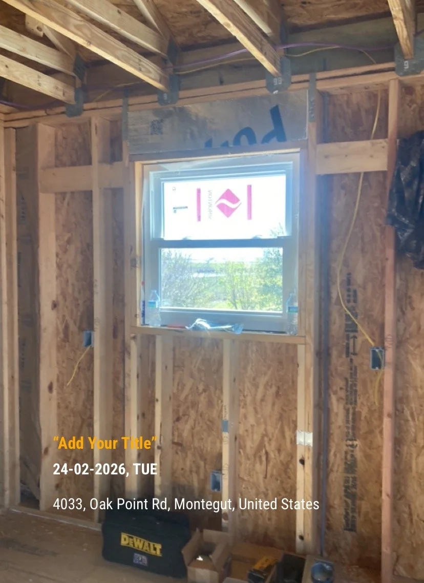 Interior of a room under construction with exposed wooden framing, electrical outlets, and a small window showing outside trees.