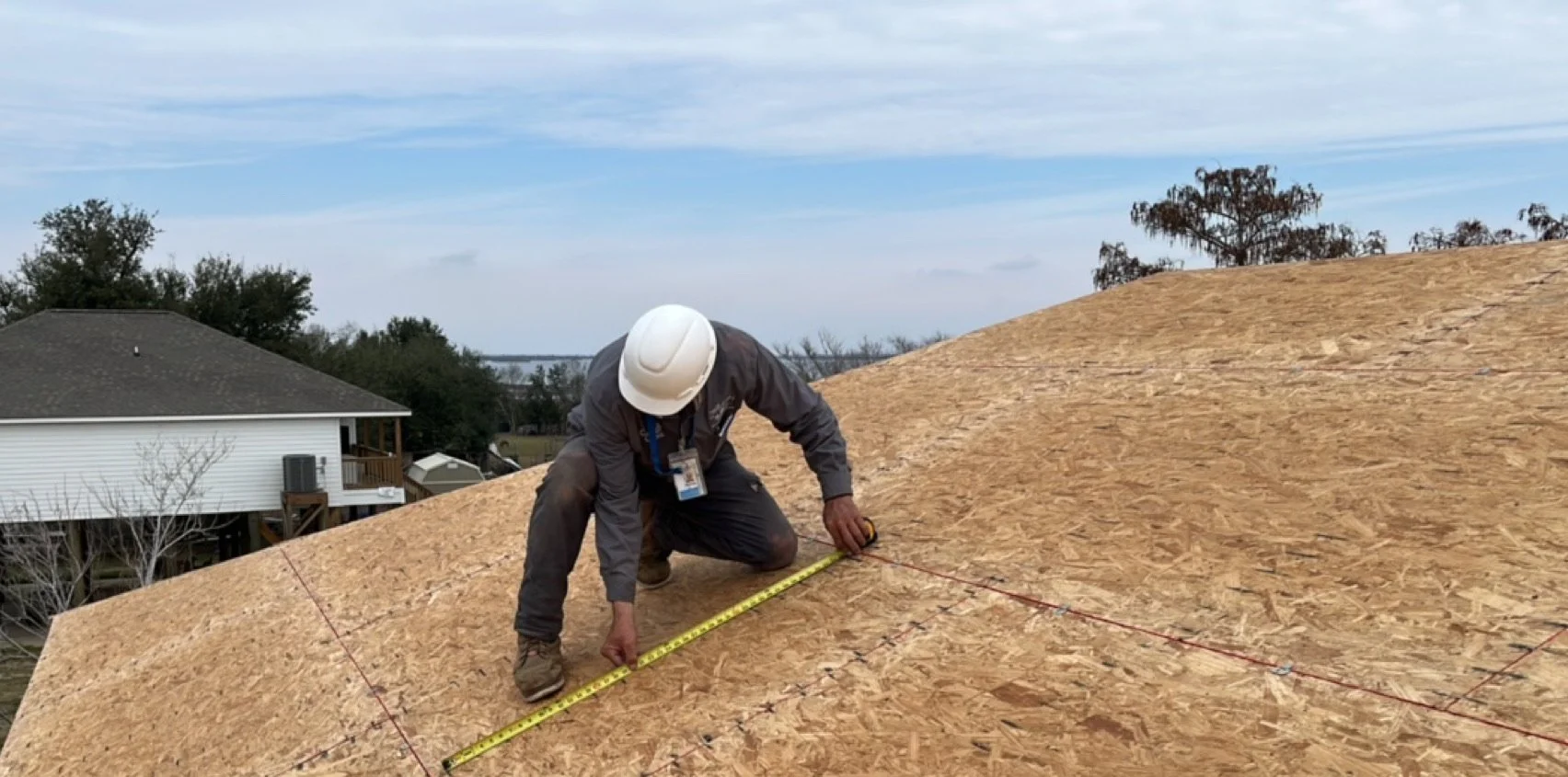A construction worker with a white helmet and gray jacket measuring a wooden surface on a roof with a yellow tape measure during daytime.