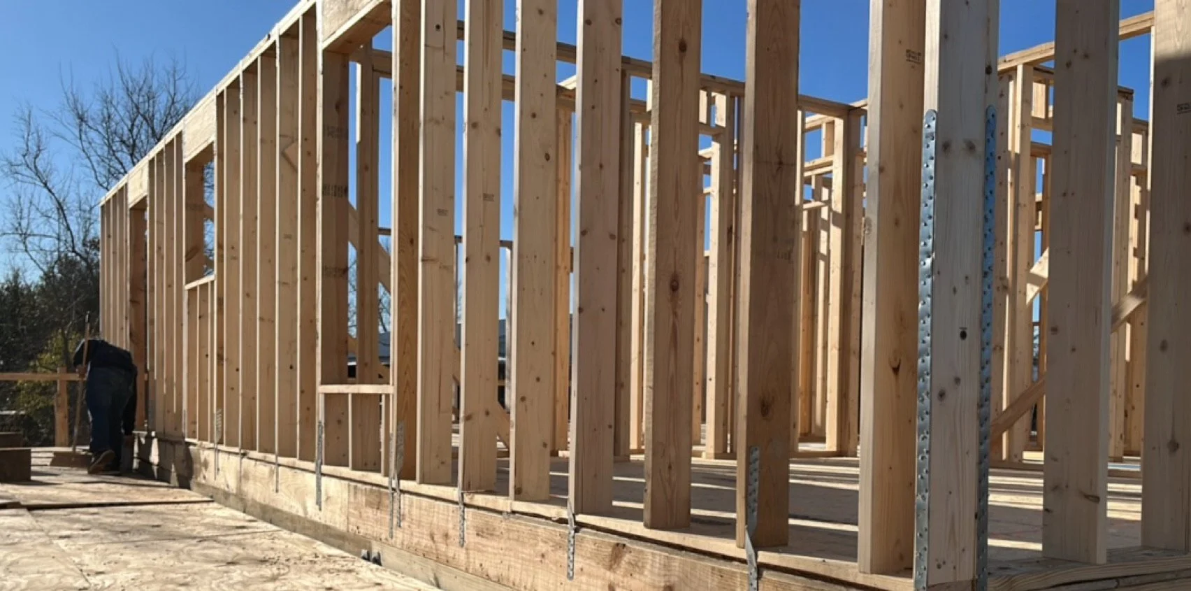 Wooden framing for a house under construction with studs and support beams, showing a clear blue sky in the background.