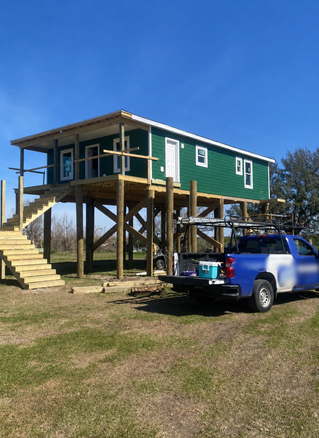 A house under construction on stilts, with a green exterior and white doors and windows. Wooden stairs lead up to the front porch, and a blue pickup truck with construction supplies is parked nearby.