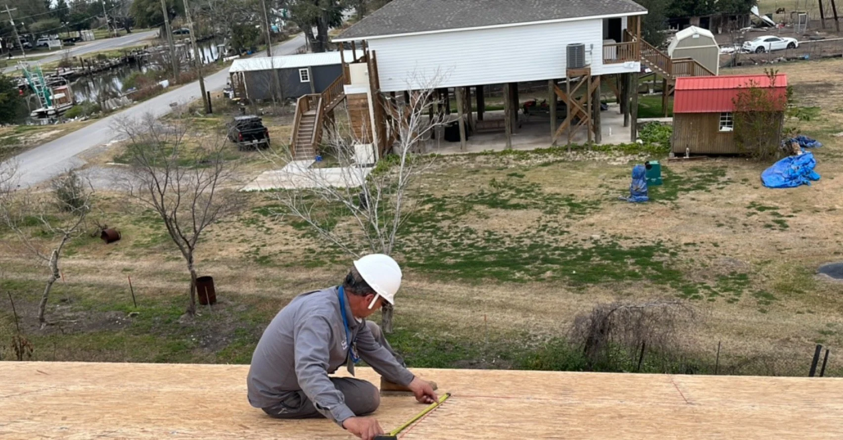 A construction worker wearing a white safety helmet and gray work clothes is measuring a wooden surface with a tape measure on a rooftop. In the background, there is a residential backyard with a large tree, small shed, and a house on stilts. The yar