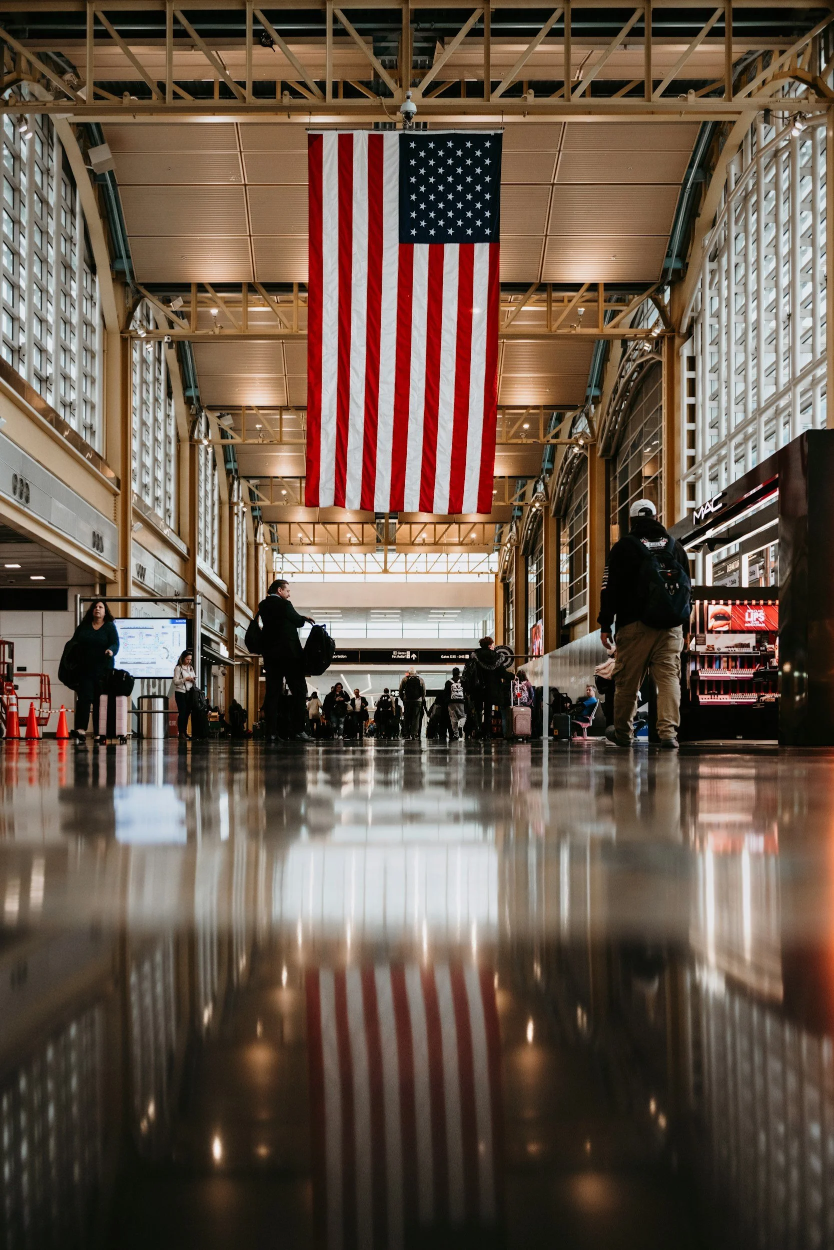 Large American flag hanging in an airport terminal with travelers walking and sitting.