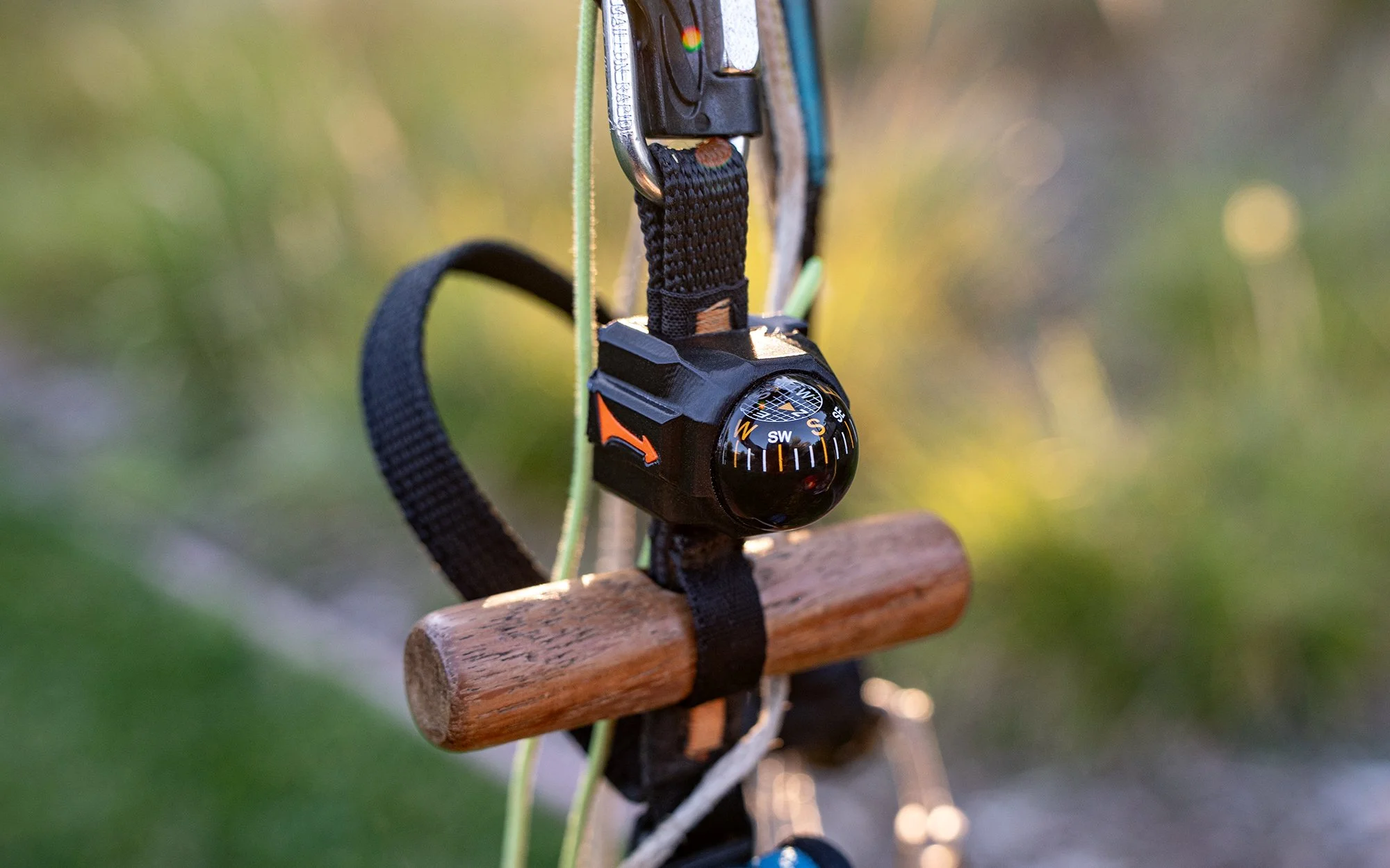 Close-up of a bluish-black compass attached to a black strap, hanging from a hook on a wooden structure with a blurred outdoor background.