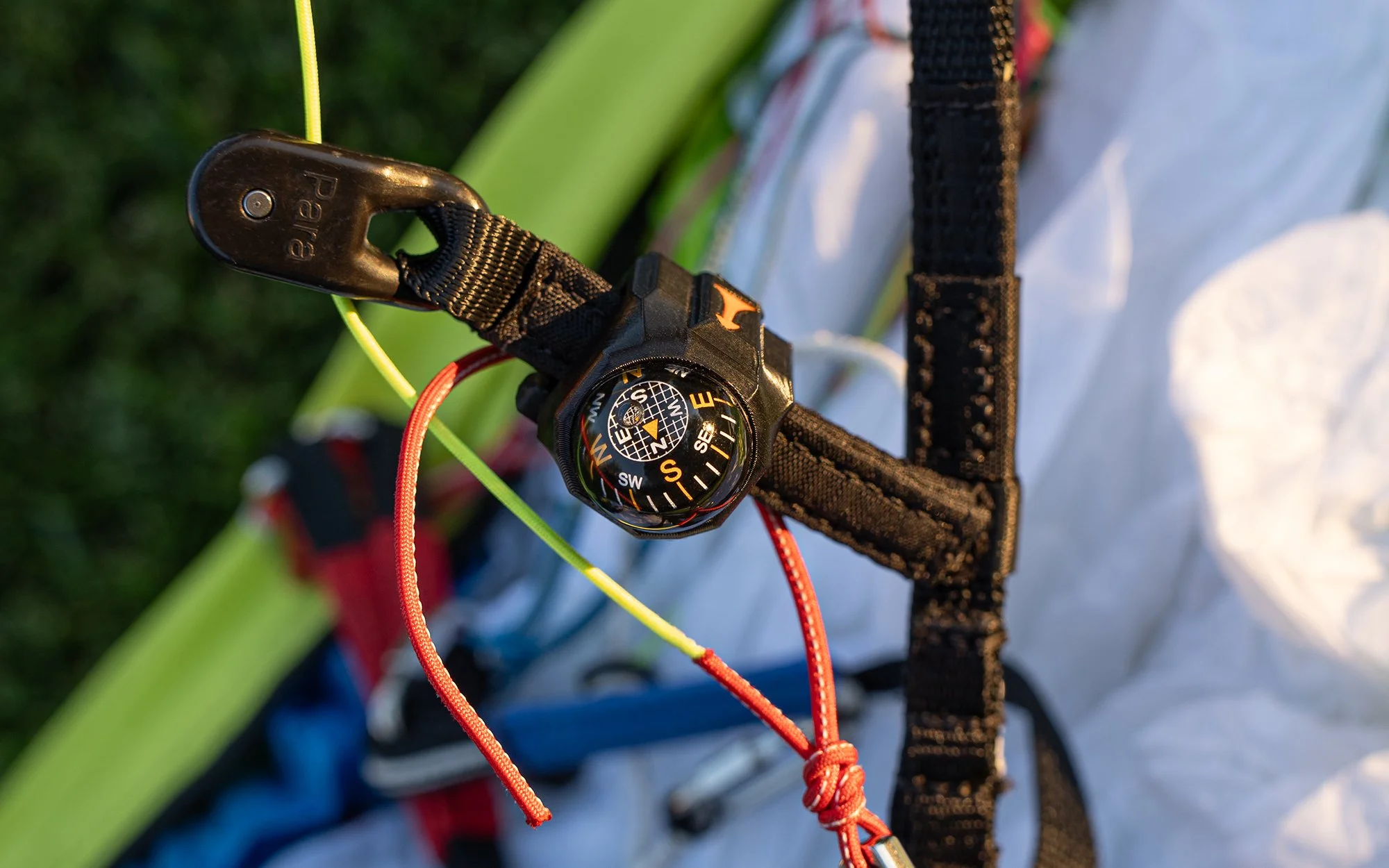 Close-up of an orange and black climbing harness with a black carabiner, a compass, and red climbing rope attached to it, on a green background.