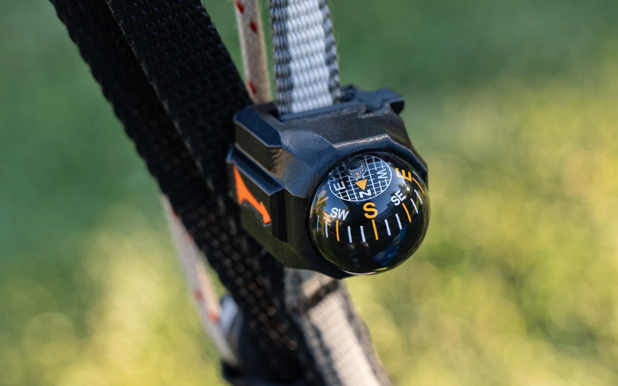 A close-up of a black compass attached to a strap, hanging from a piece of equipment or gear outdoors with blurred green background.