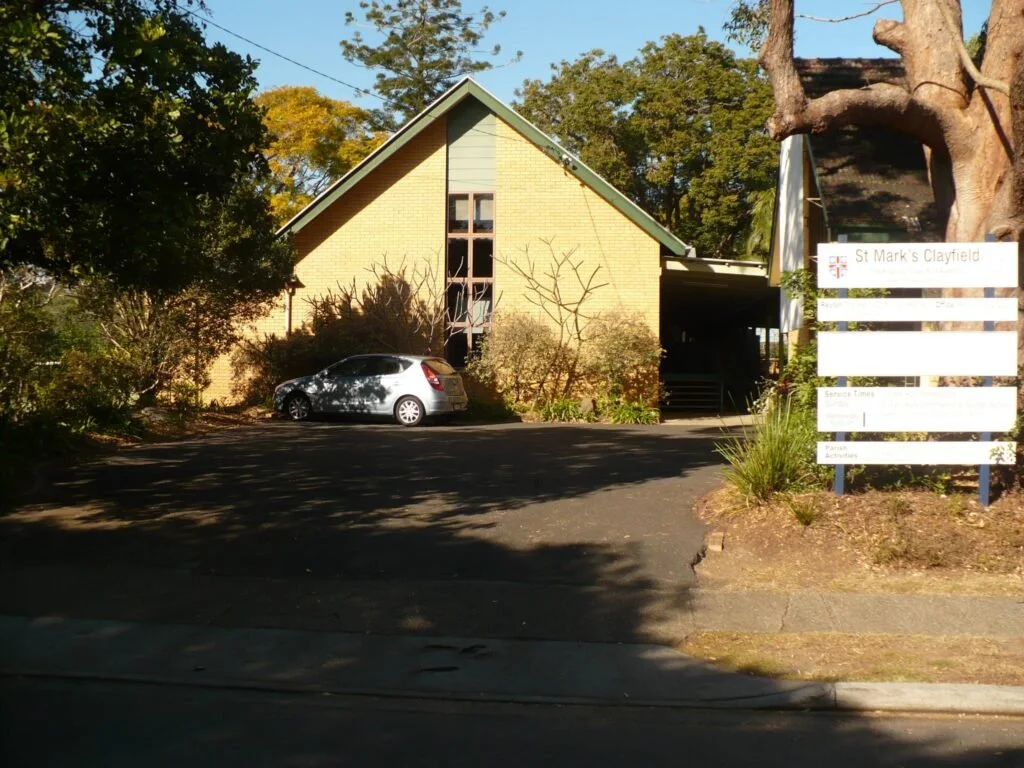 A brick church with a pointed roof and large front window, surrounded by trees and bushes, with a parking area and a white car in front, and a sign indicating the church's name as St. Mark's Clayfield.