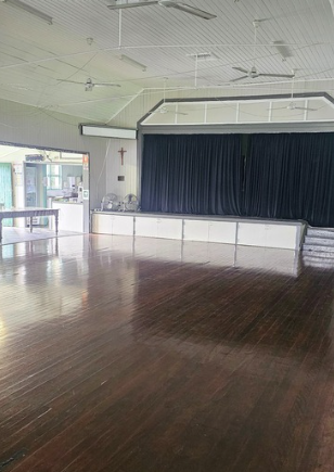 Empty indoor space with wooden floor, stage with black curtains, ceiling fans, and a cross on the wall.