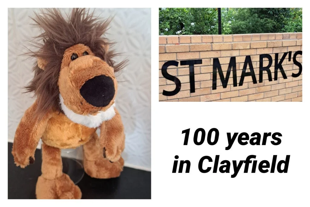 A plush lion toy with a brown mane, a large black nose, and a white patch on its chest placed on a black surface against a textured white background. A brick wall with the words "ST MARK'S" in black letters and a green tree are visible in the background, with text indicating a 100-year celebration at Clayfield.