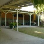 A covered outdoor patio with white support poles, surrounded by green grass and a few potted plants.