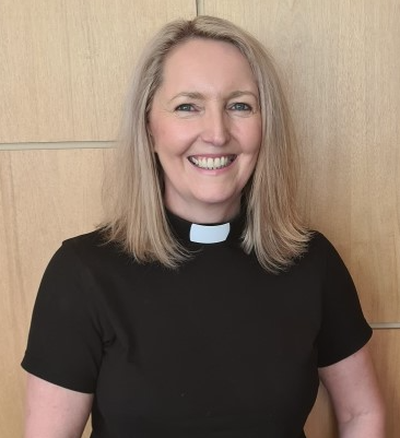 A woman wearing a black clergy shirt with a white clerical collar, smiling in front of a wooden background.