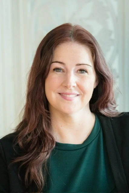 A woman with long brown hair and fair skin, smiling, wearing a dark green top and a black cardigan, sitting indoors in front of a light-colored background.