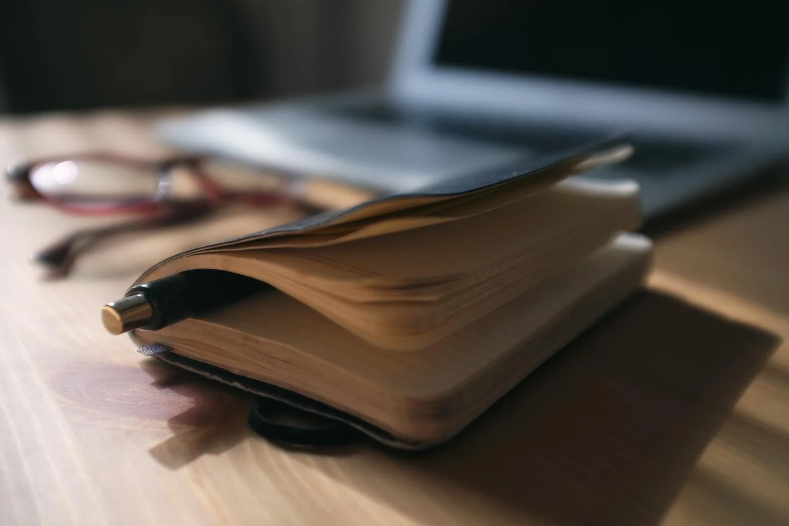 Open notebook with lined pages and a black pen resting on it, blurred eyeglasses and a laptop in the background.