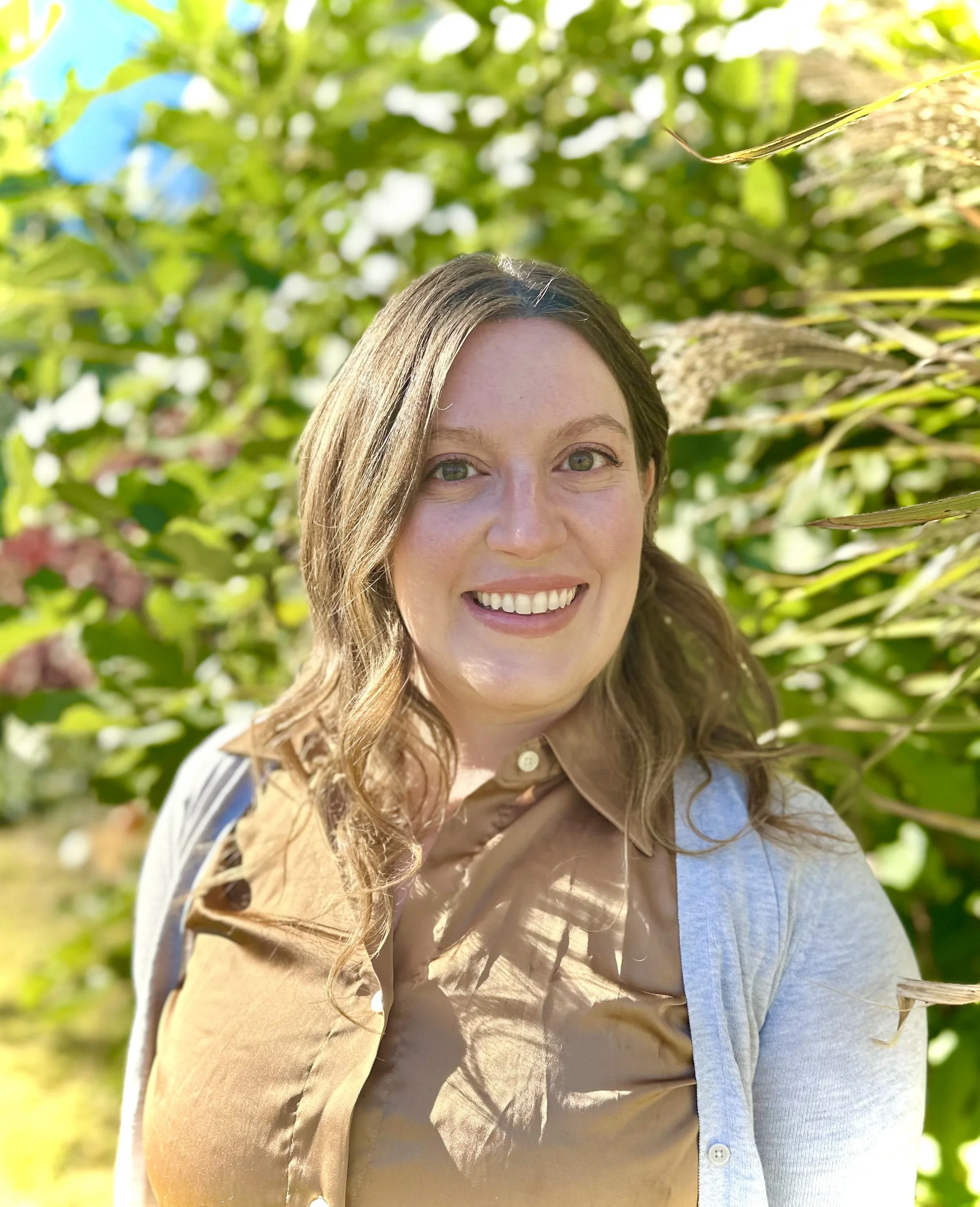 Close-up of a smiling woman with wavy brown hair, wearing a light blue shirt, standing in a green outdoor setting with sunlight filtering through the leaves.