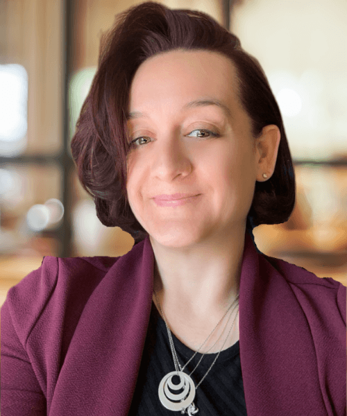 A woman with shoulder-length dark hair, wearing a purple blazer and a black top, smiling slightly in an indoor setting with blurred background.