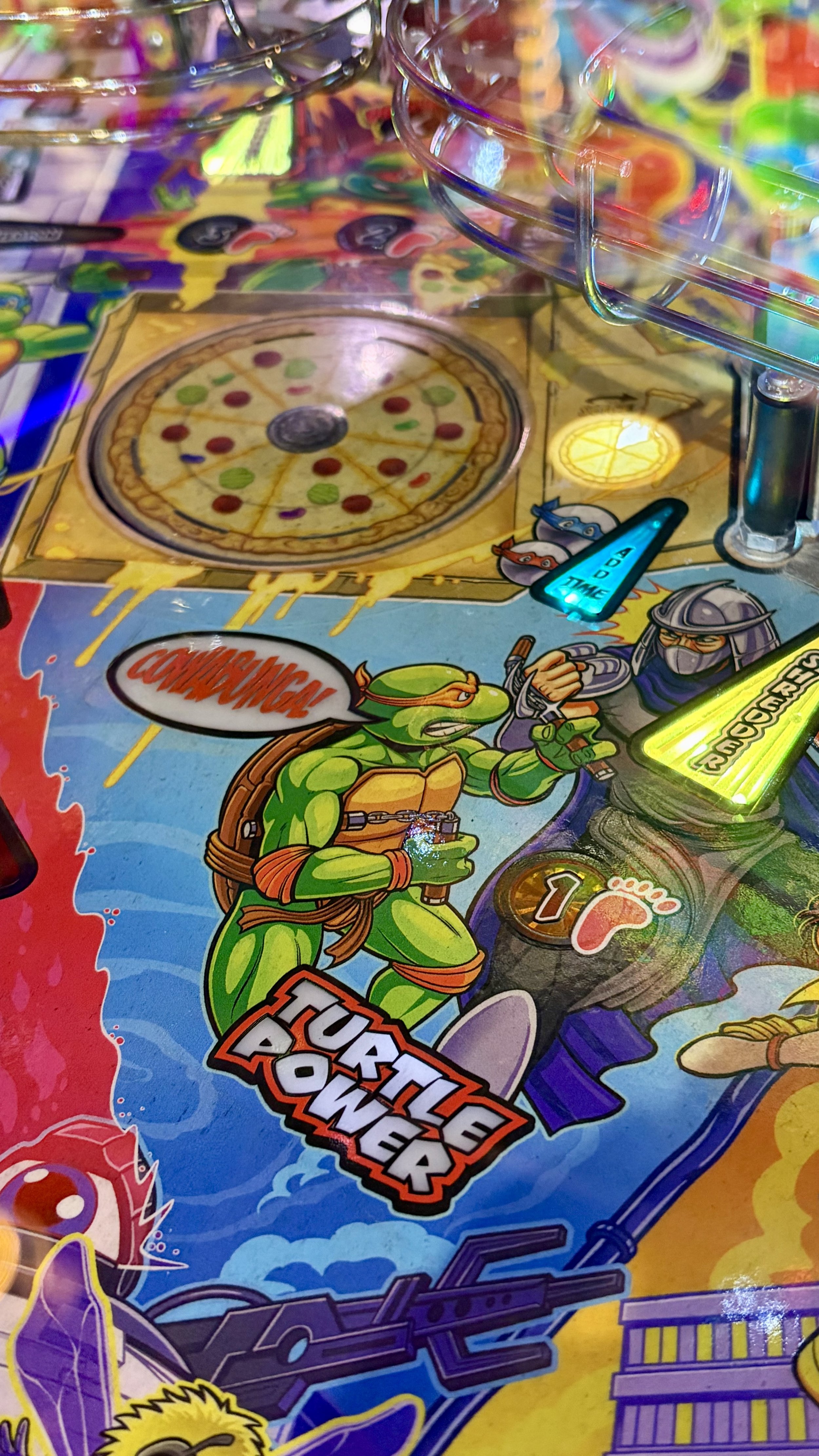 Close-up of a Teenage Mutant Ninja Turtles-themed pinball machine, featuring a pizza with musical notes and game characters, with colorful lights and artwork.
