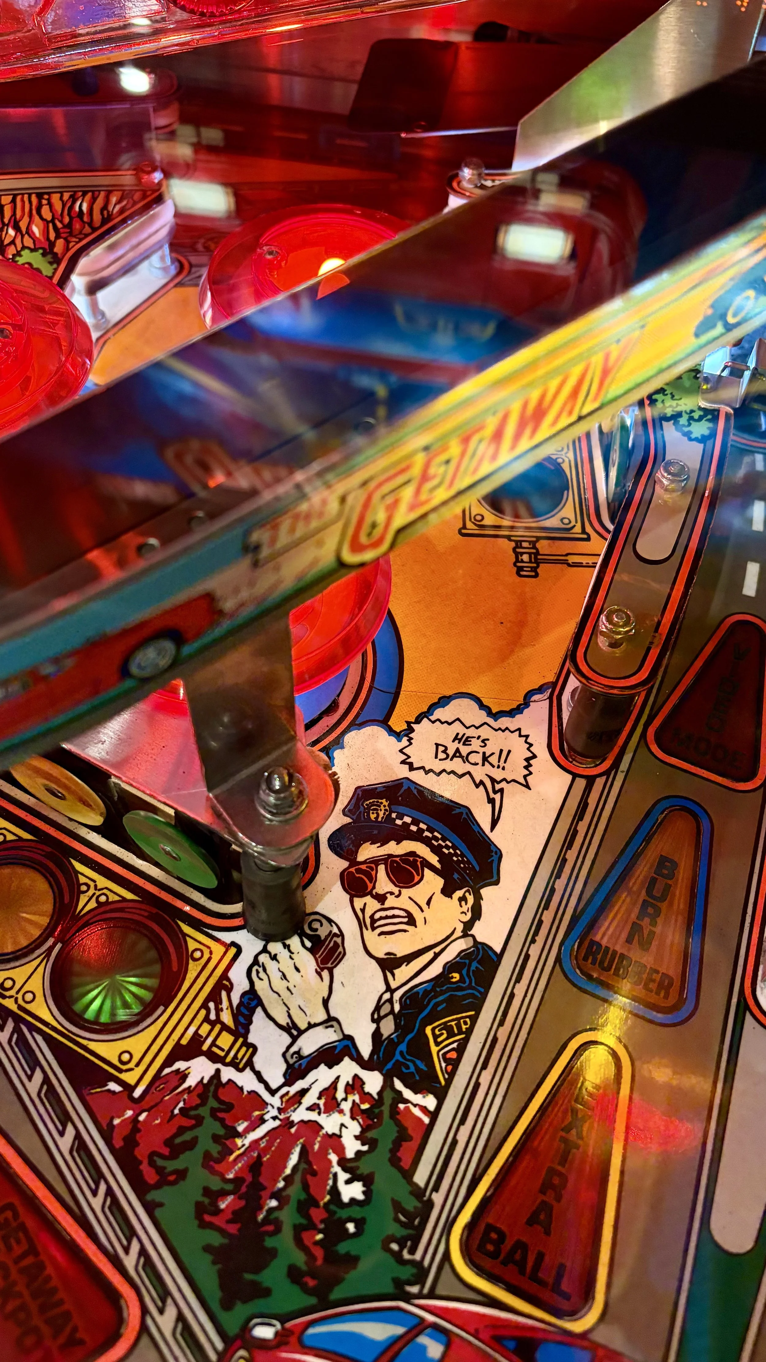 Close-up of the game Getaway a vintage pinball machine playfield with flashing lights, artwork, and the phrase "He's BACK!!" with a cartoon police officer.