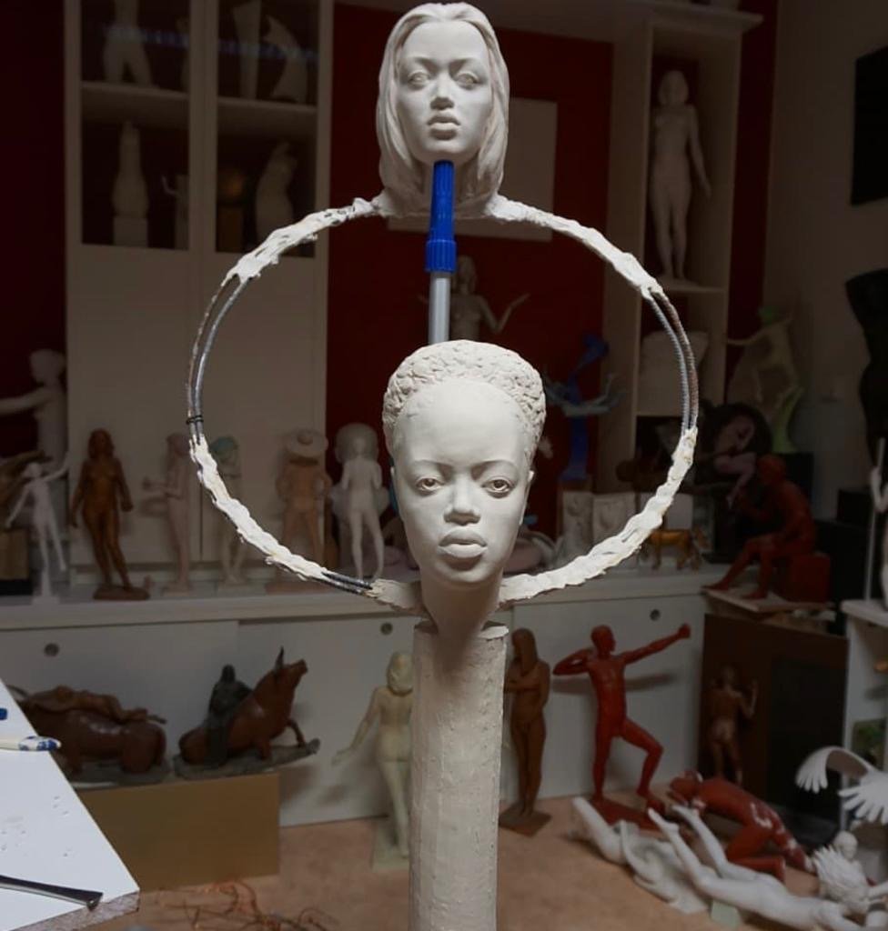 A sculpture of two female faces, one larger and one smaller, connected by a circular ring, in an art studio surrounded by various other sculptures.