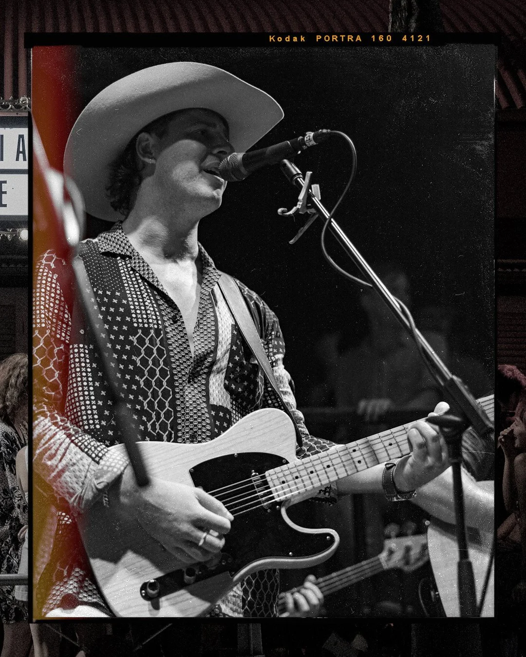 Black and white photo of a Nick Trovas wearing a cowboy hat and patterned shirt, singing into a microphone while playing an electric guitar.