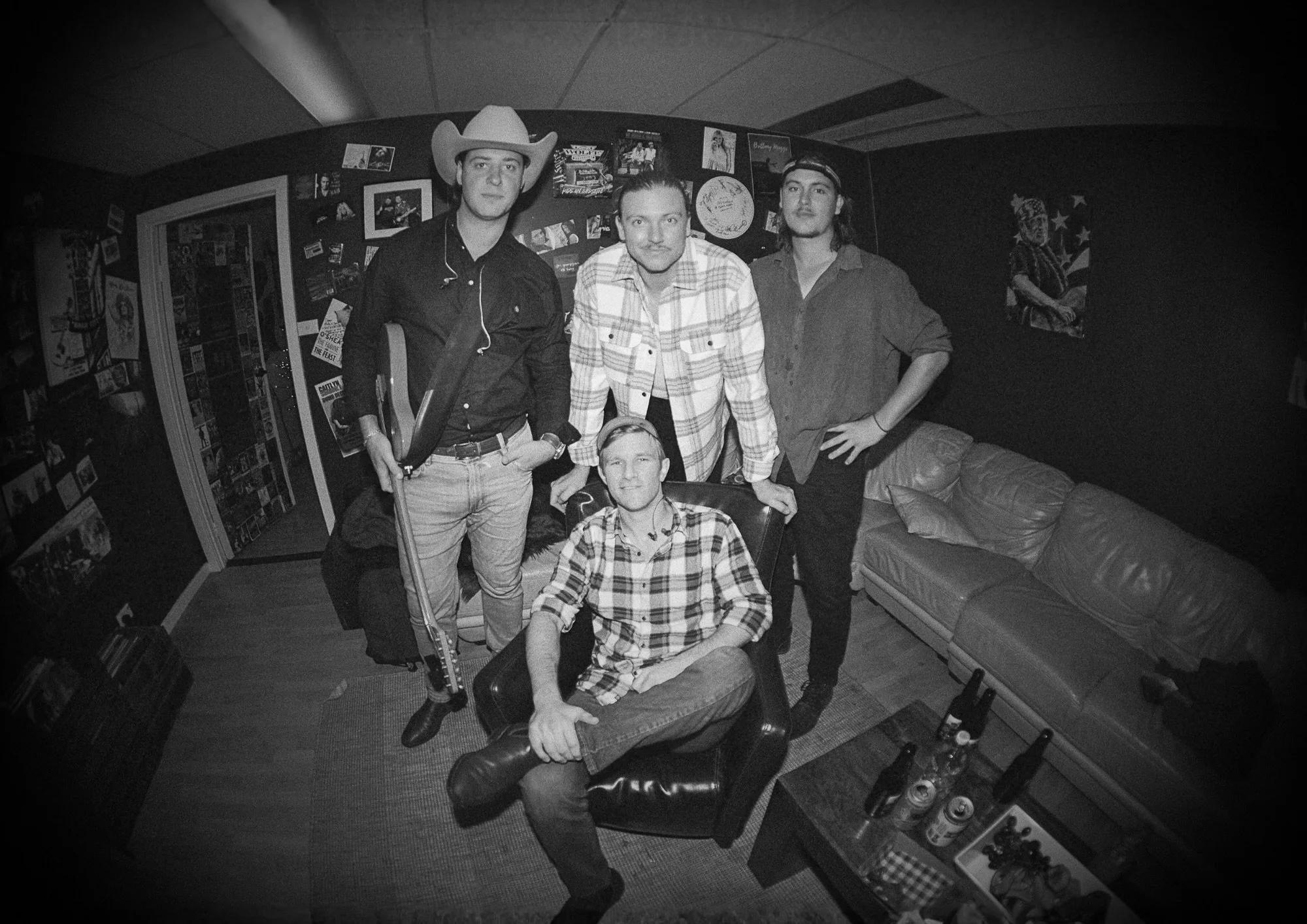 Four men in a room with posters on the wall, one wearing a cowboy hat and holding a guitar, another standing with his hand on his hip, and two sitting on chairs, all facing the camera.