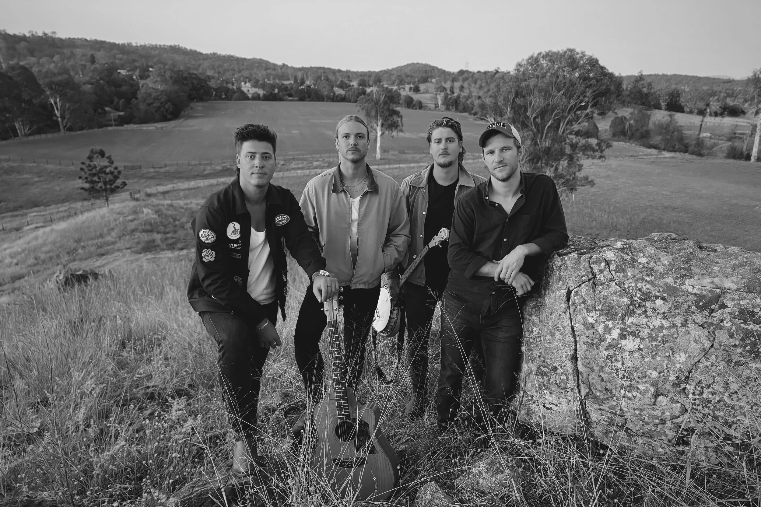 Four young men standing outdoors in a grassy field with trees and rolling hills in the background. One of them is holding an acoustic guitar, and another has a helmet in hand. The picture is in black and white.