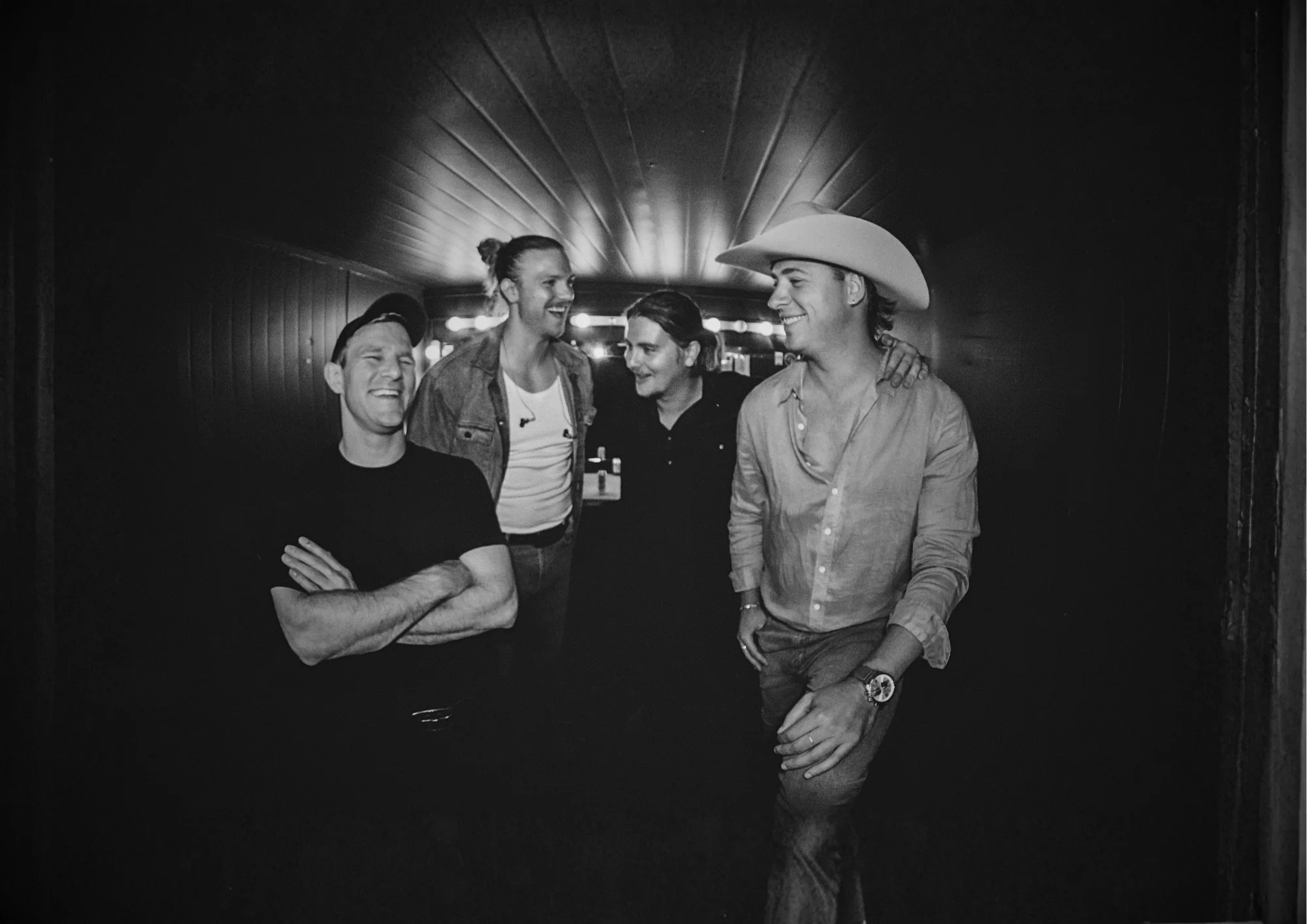 Four friends are laughing and smiling together in a corridor with a domed ceiling, some wearing hats and casual clothing.