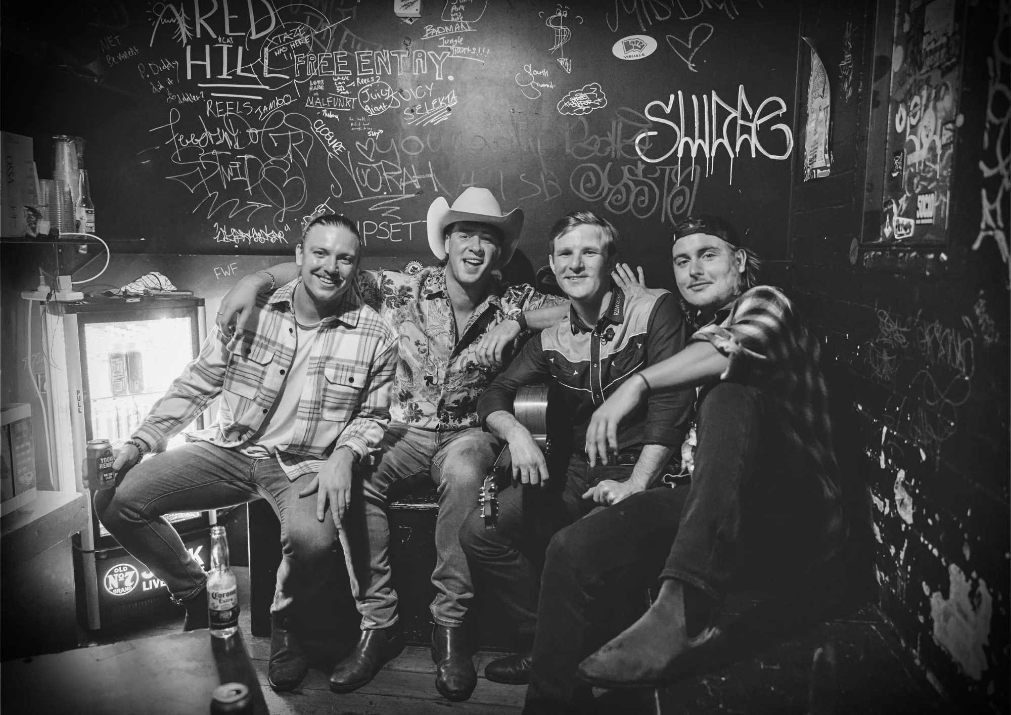 Four men sitting closely together in a bar, smiling at the camera. One man is wearing a cowboy hat, and another has a baseball cap backwards. There are graffiti and artwork on the black wall behind them, and a small fridge with drinks and a bottle of
