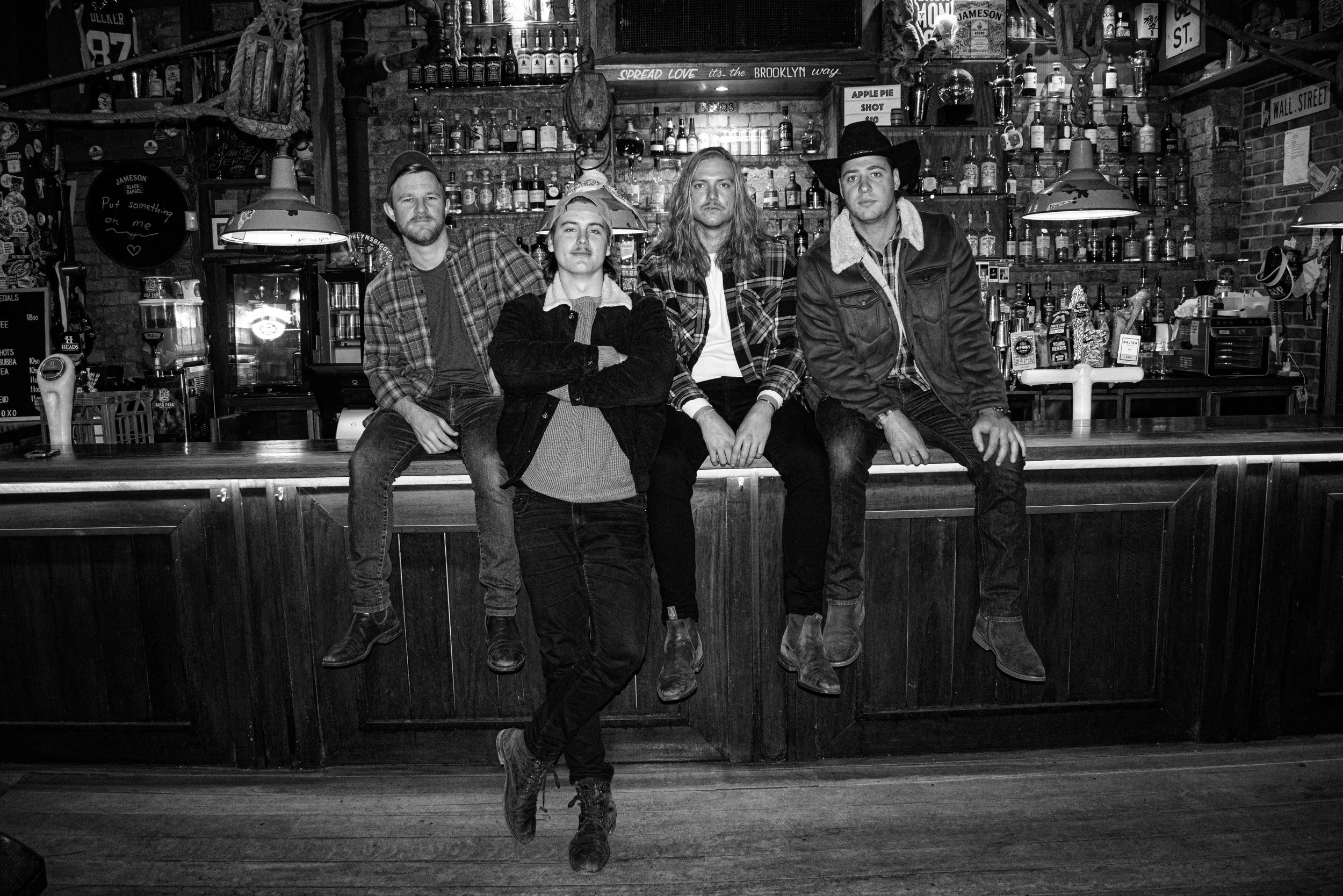 Four young men in The Wet Whistles, posing together behind a bar counter in a rustic, eclectic bar setting with shelves of liquor bottles and bar equipment in the background.