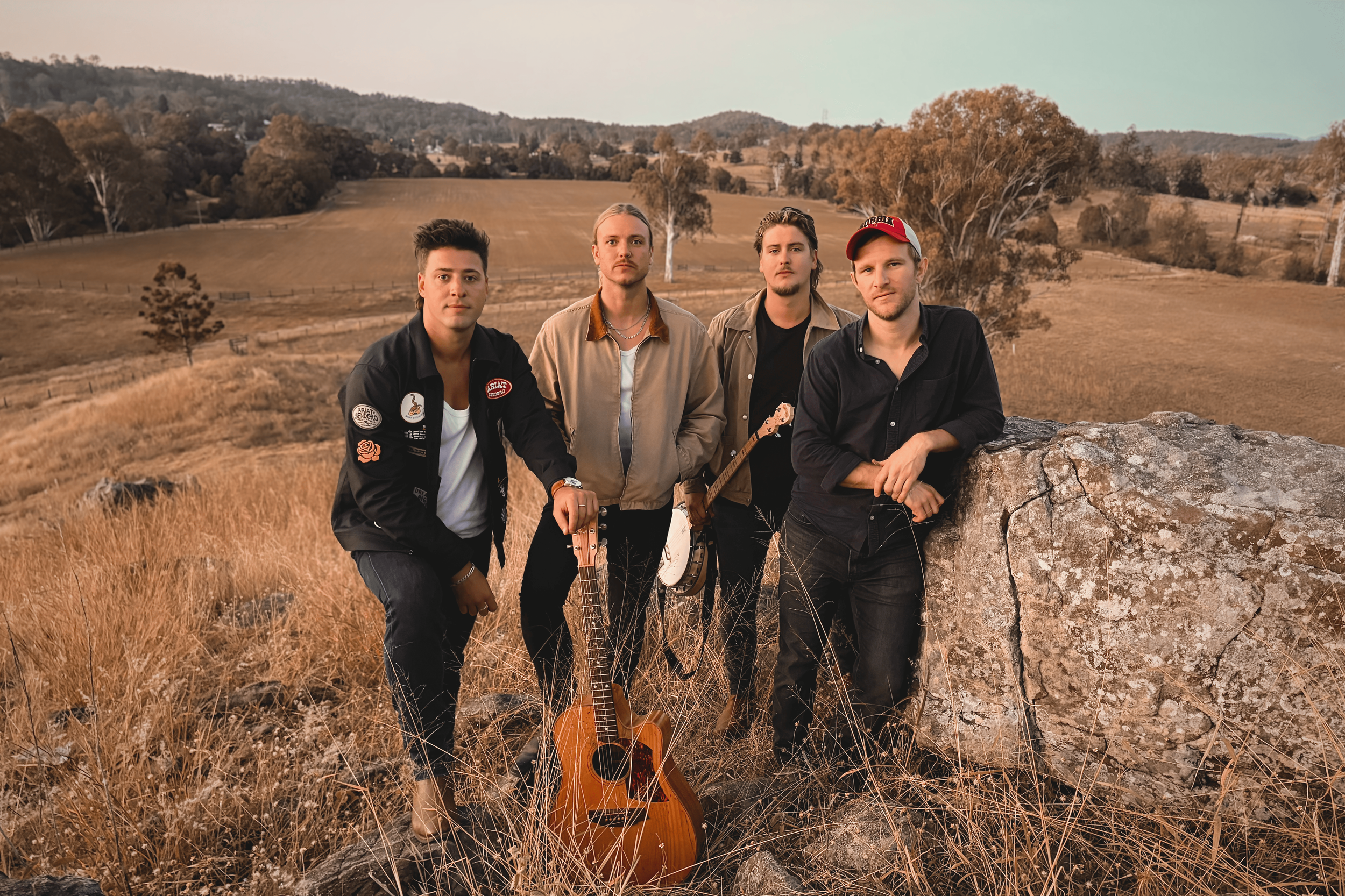 Four young men standing outdoors in a grassy field with rolling hills and trees in the background, one holding a guitar, with another guitar leaning against a rock in front of them.