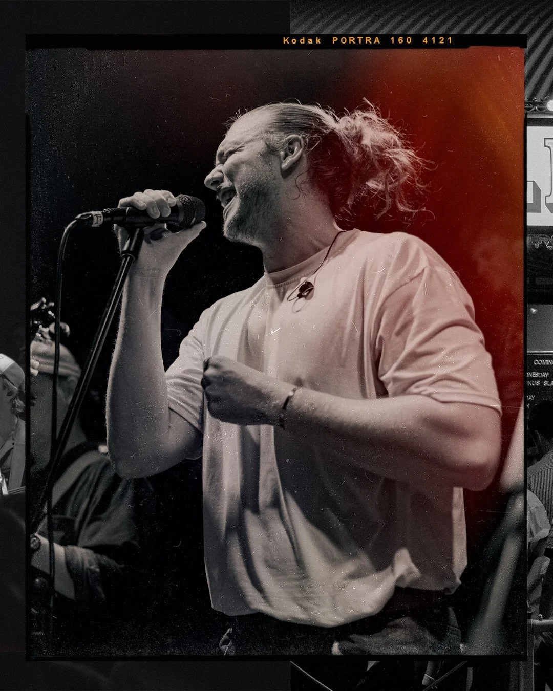 A black and white photo of a Jake Hill singing into a microphone, with long hair tied back, wearing a casual shirt, and passionately performing.