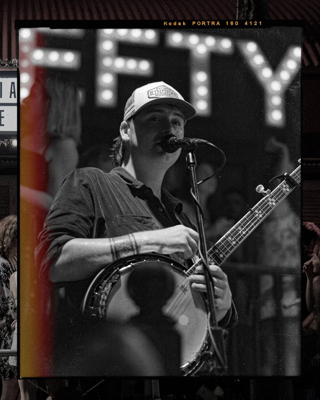 Black and white photograph of musician, Jordan Hill, singing into a microphone while holding a banjo and a guitar, wearing a baseball cap with a patch that says 'Ringer Western,' behind a lit marquee sign.
