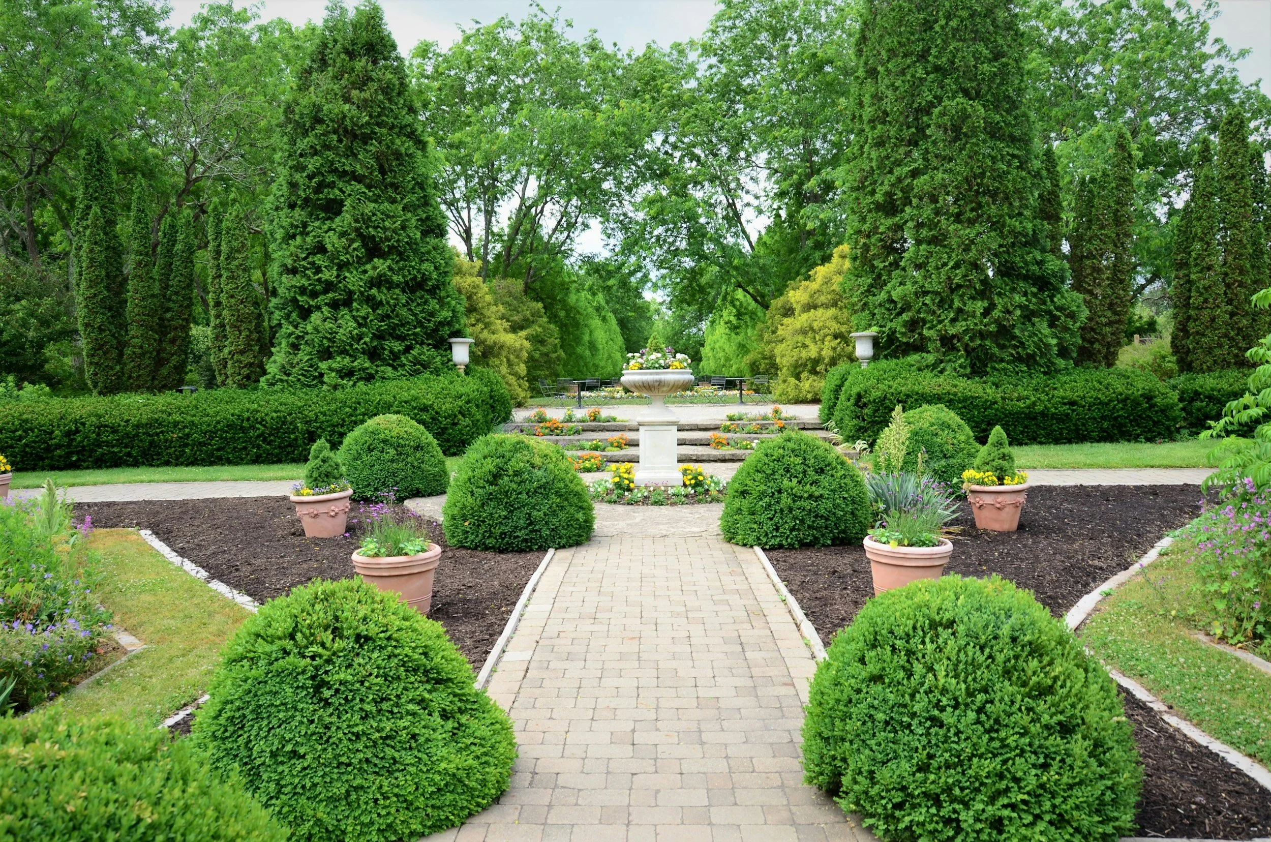 A lush, well-maintained garden with a paved path, decorative potted plants, neatly trimmed bushes, a central flower fountain, and tall green trees in the background.