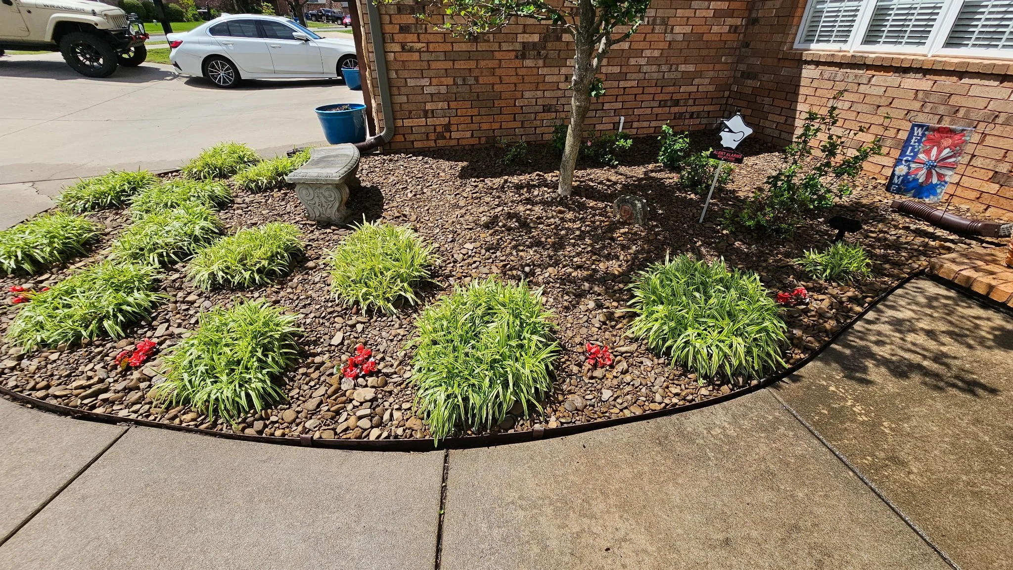 A small landscaped flower bed bordered with black edging, containing green plants with yellow and red flowers, a decorative stone bench, a young tree, and various signs and decorations, located next to a brick house wall and a concrete sidewalk.