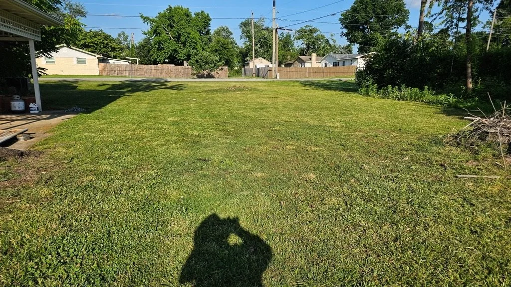A backyard with a well-maintained lawn, a small covered patio area on the left, and trees lining the yard's perimeter. There are houses and utility poles in the background, under a clear blue sky with some clouds.