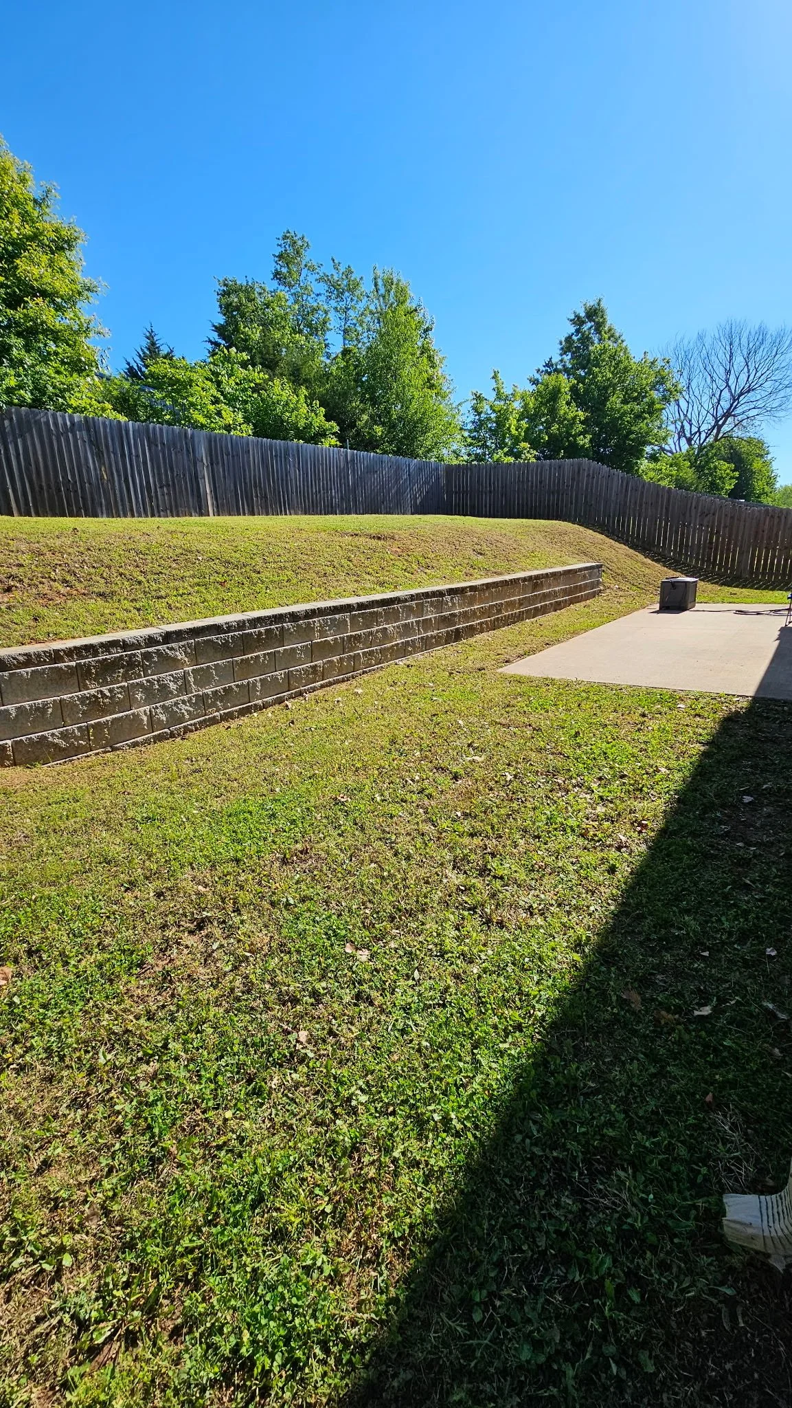 Backyard with a grass lawn, a stone retaining wall, a concrete patio, a wooden fence, and trees against a blue sky.
