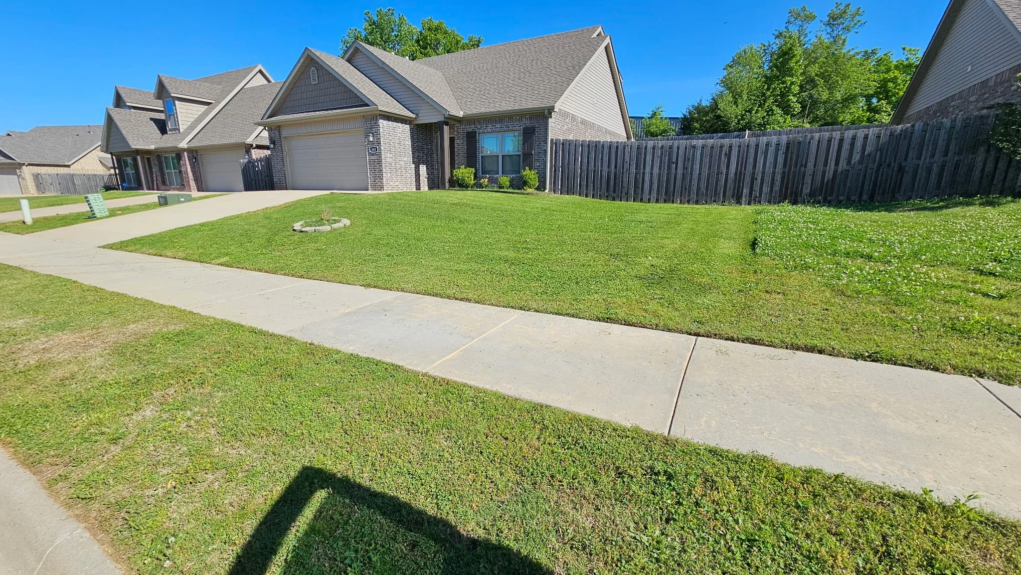 Front yard with freshly mowed grass, sidewalk, and houses with garages in a suburban neighborhood under a clear blue sky.