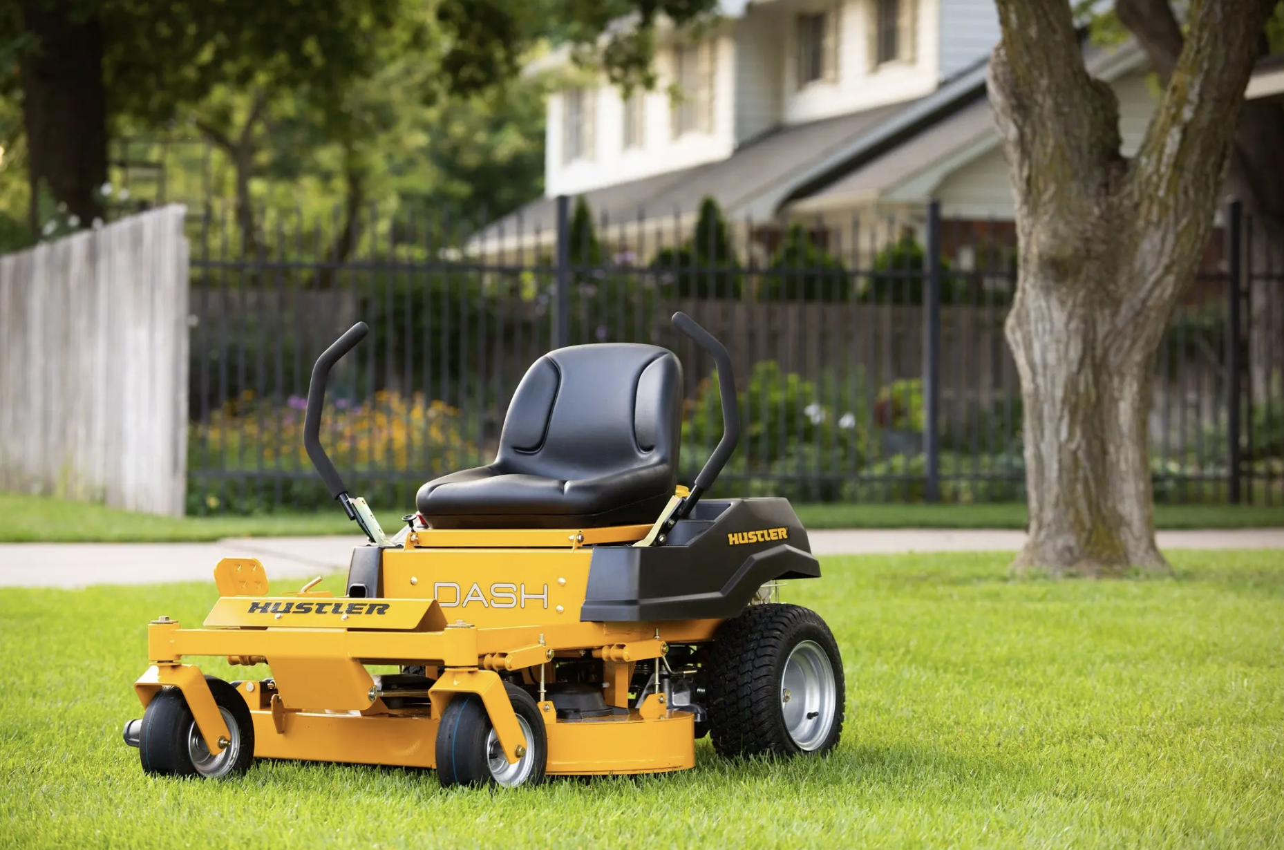 A yellow Hustler Dash riding lawn mower on a lush green lawn in a residential backyard, with trees, a black metal fence, and a house with white siding and a gray roof in the background.