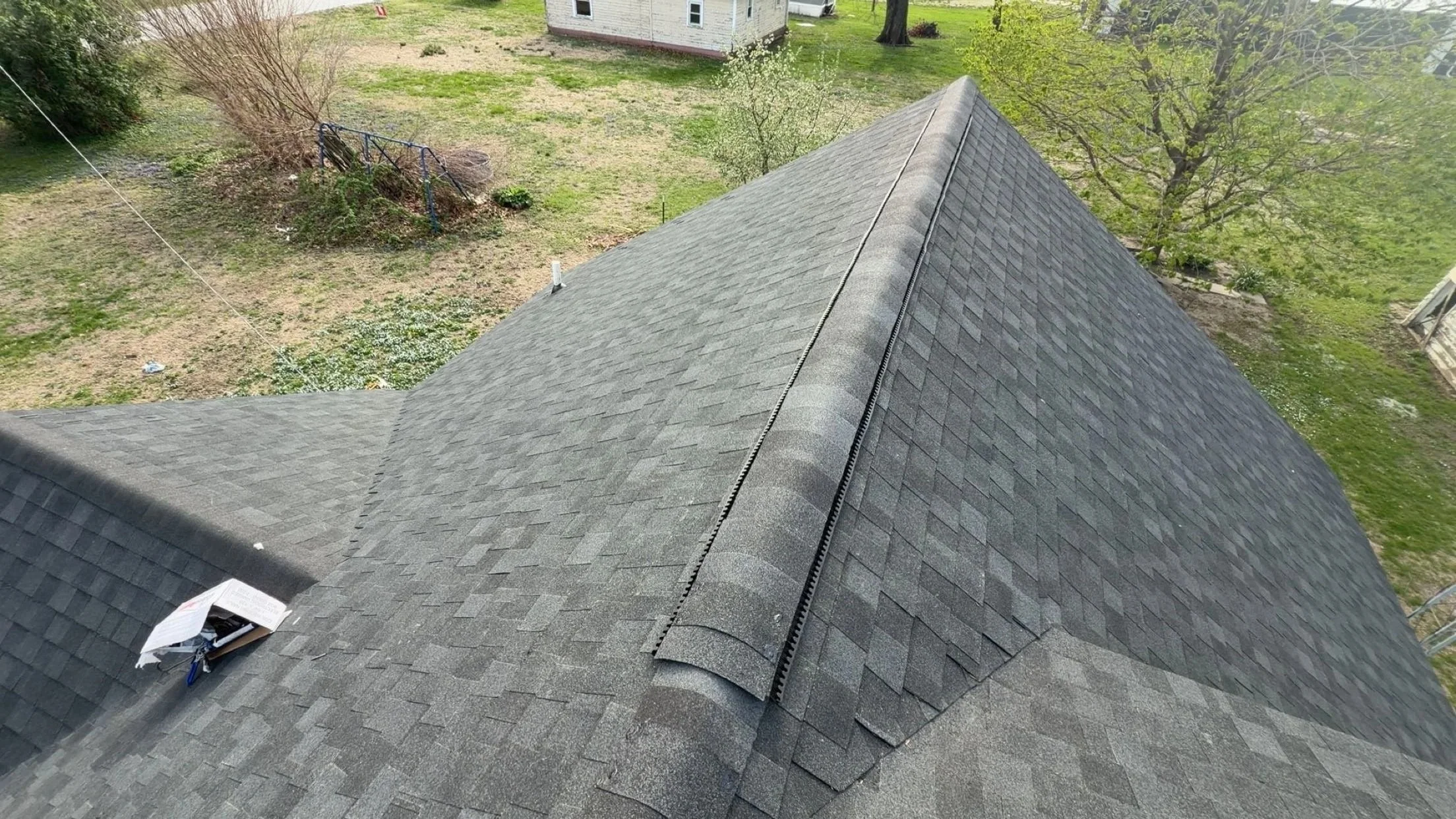 Aerial view of a gray shingled house roof with a chicken ladder along the seam, a small open box with tools or materials on the roof, and a backyard with grass, trees, a bush, a clothesline, and a small structure in the distance.