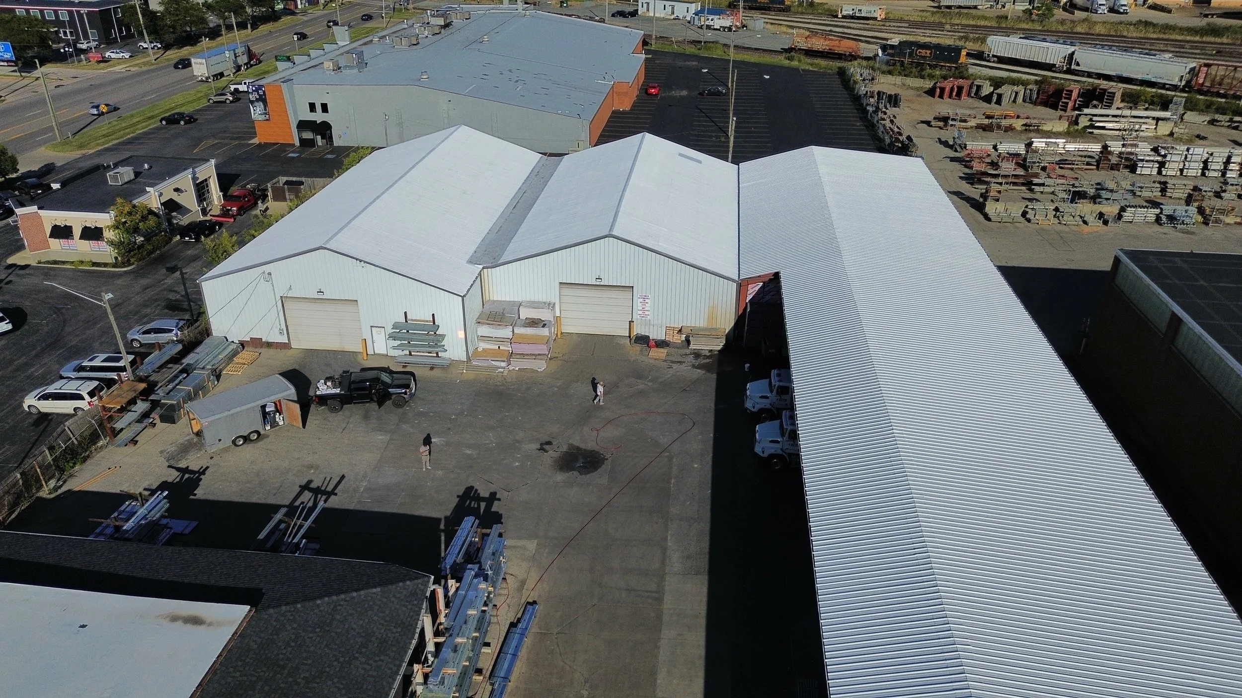 Aerial view of an industrial warehouse and storage yard with construction materials, vehicles, and adjacent parking lot near train tracks.