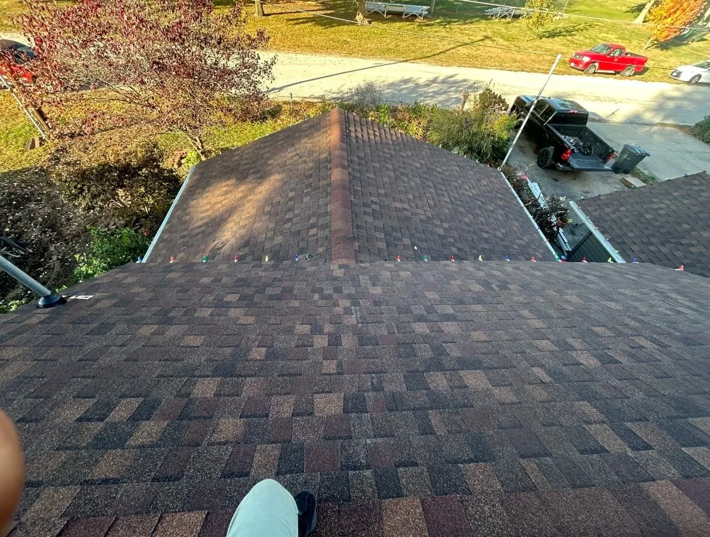 View from the roof of a house looking down at neighboring rooftops, trees, parked pickup trucks, and a street with parked cars.